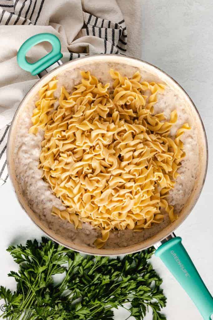 Cooked egg noodles on top of ground beef mixture in a skillet.