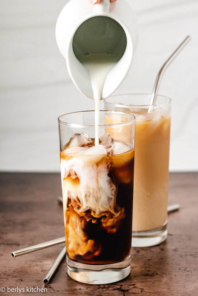 Coffee creamer being poured into a glass of iced coffee.