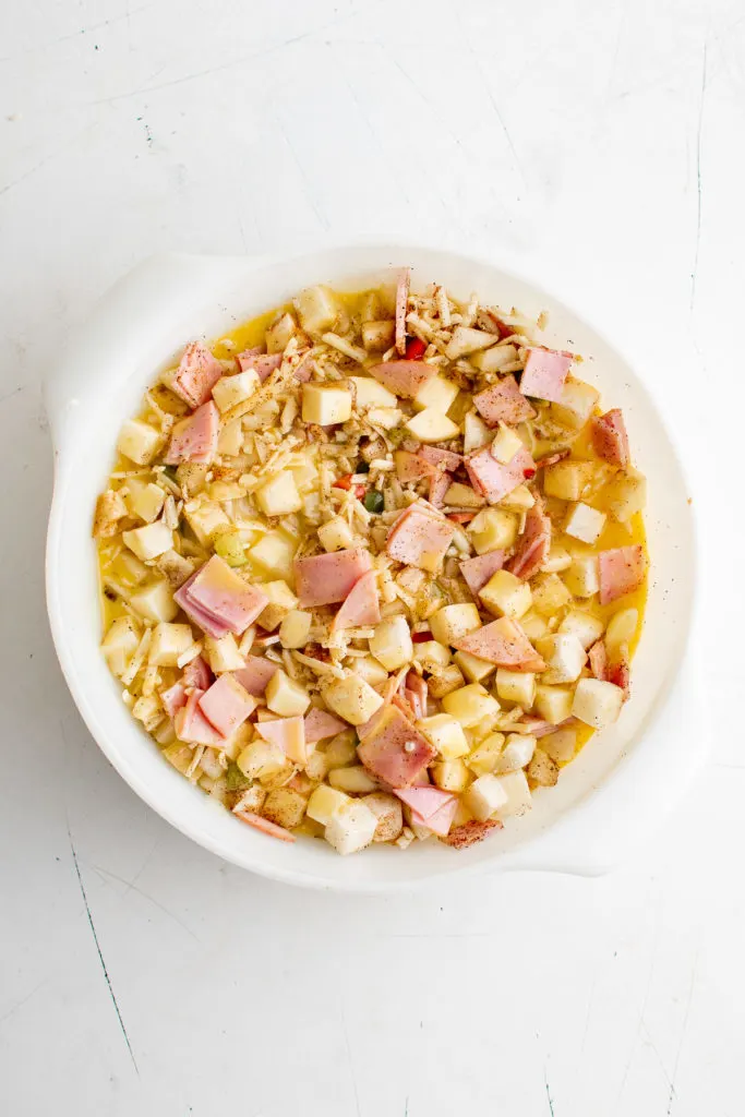 Eggs added to potatoes, cheese, Canadian bacon, salt, pepper and chili powder in a dish.