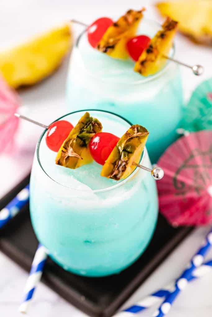 Cherries and pineapple on top of a glass of blue hawaiian drink.