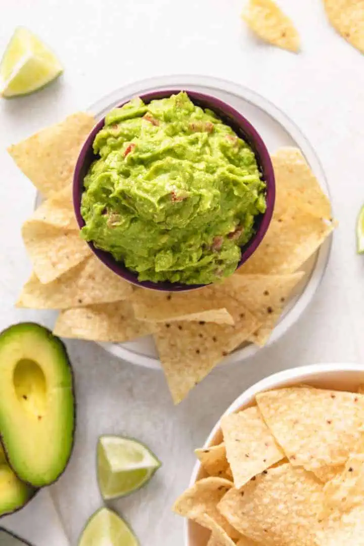 Top down view of a purple bowl of guacamole and chips, lime, and avocados.