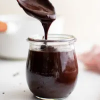 Spoon holding of hot chocolate fudge from a glass jar.