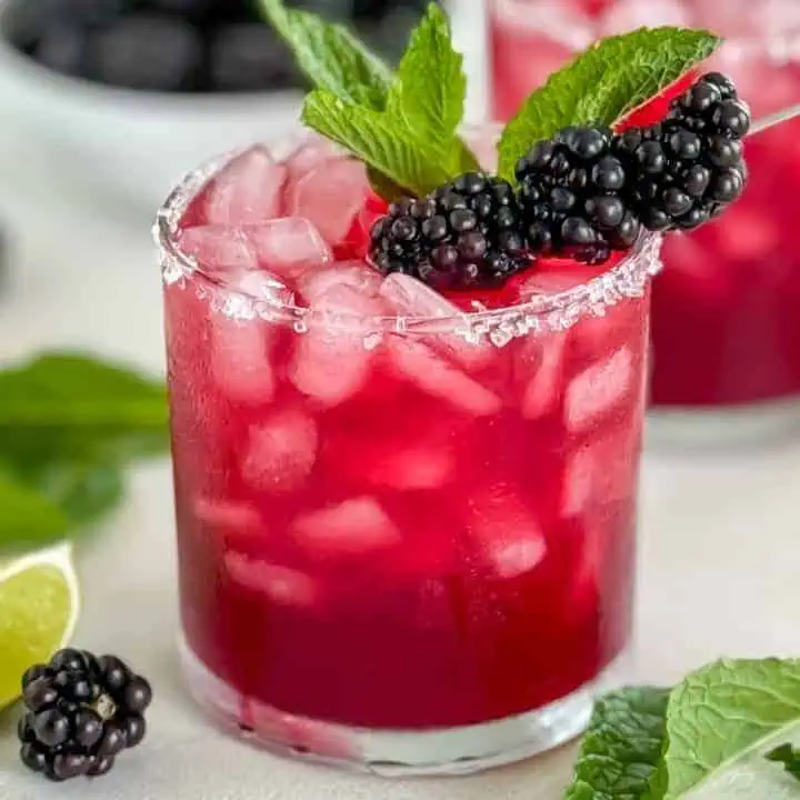 Featured image of a Blackberry Margarita in a glass with a sugared rim, garnished with fresh blackberries and mint leaves, with lime wedges and blackberries in the background.