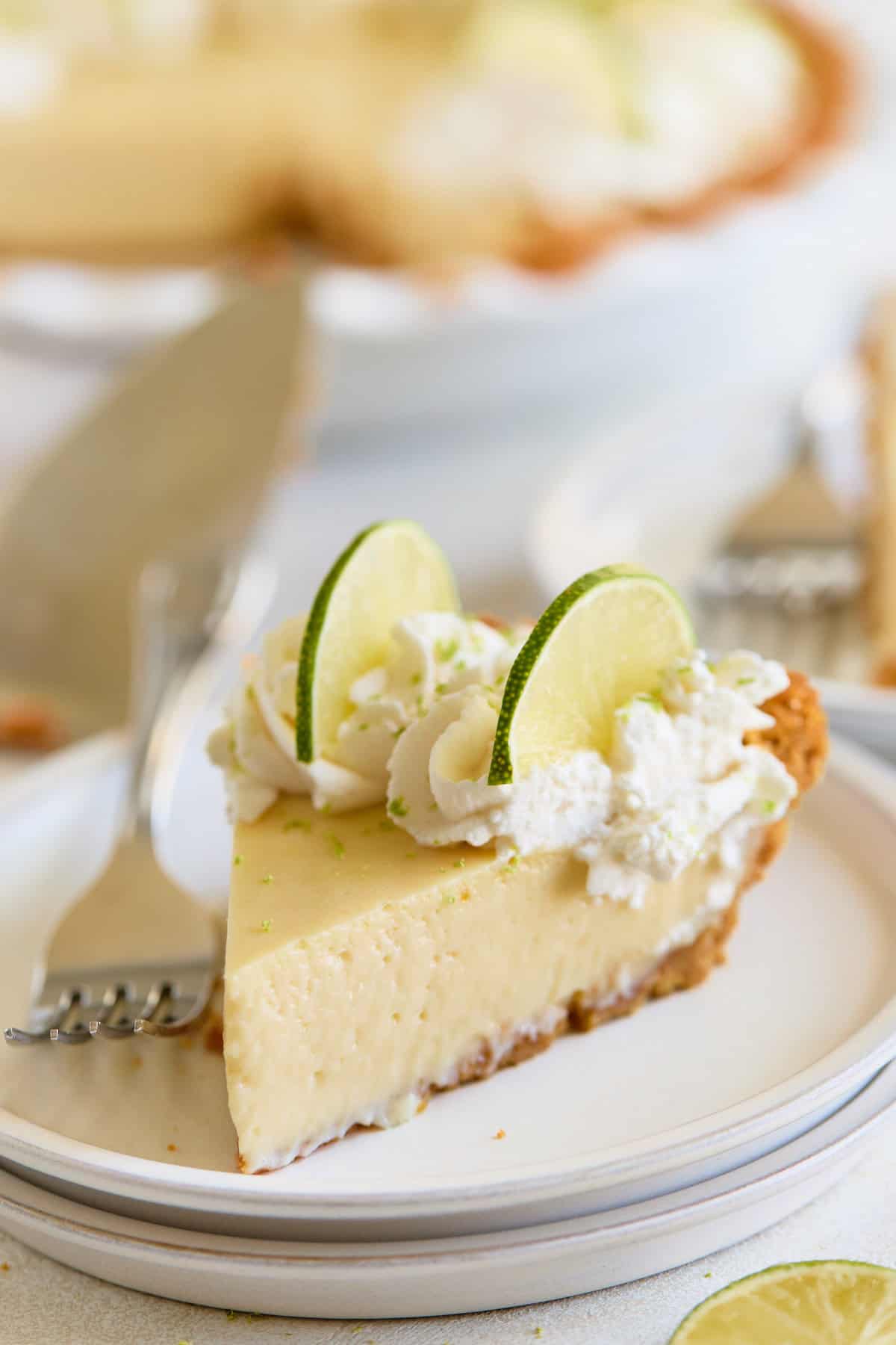 Key Lime Pie A close-up slice of creamy pie topped with whipped cream and lime slices on a white plate, with the whole pie in the background.
