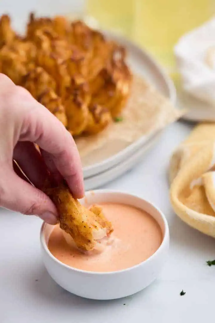 Hand dipping a piece of air fried blooming onion into a creamy dipping sauce.