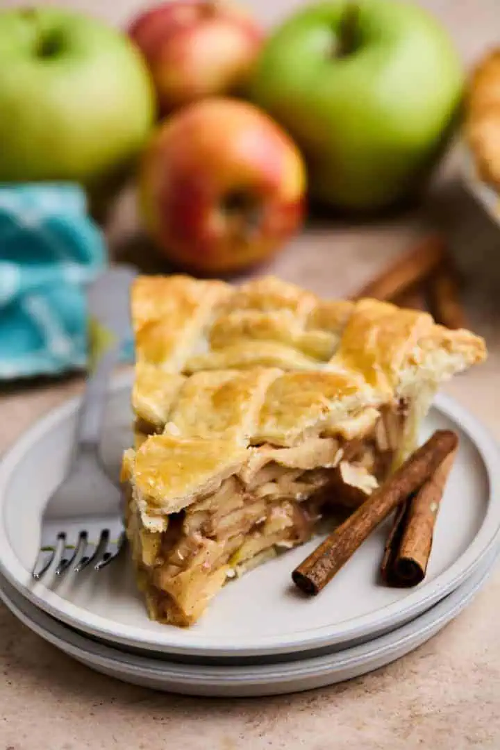 A slice of apple pie on a white plate, with cinnamon sticks beside it and whole apples in the background.