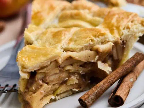A close-up slice of apple pie with a golden crust, showcasing the layered apple filling, with cinnamon sticks and a fork beside it.