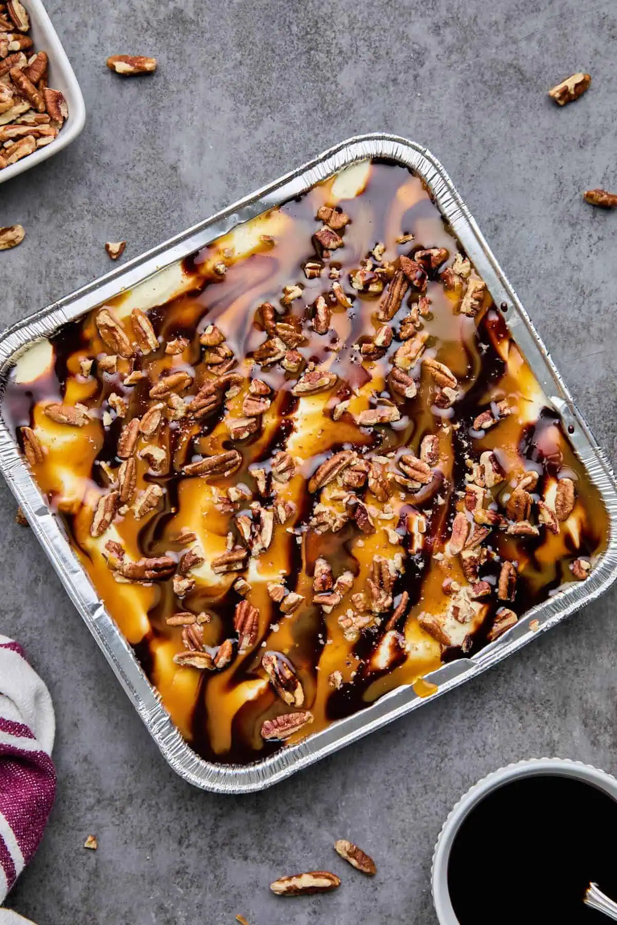 Fully assembled turtle cheesecake with chocolate and caramel sauce drizzles and pecans in an aluminum pan.