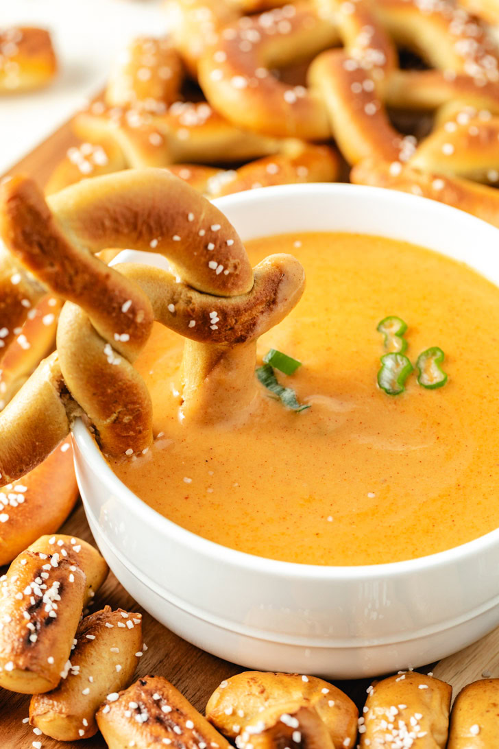 A twisted soft pretzel being dipped into a bowl of cheese dip.