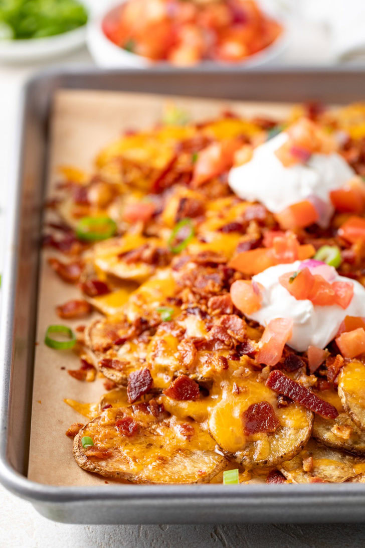 A close-up of crispy baked potato nachos topped with sour cream, diced tomatoes, and bacon.