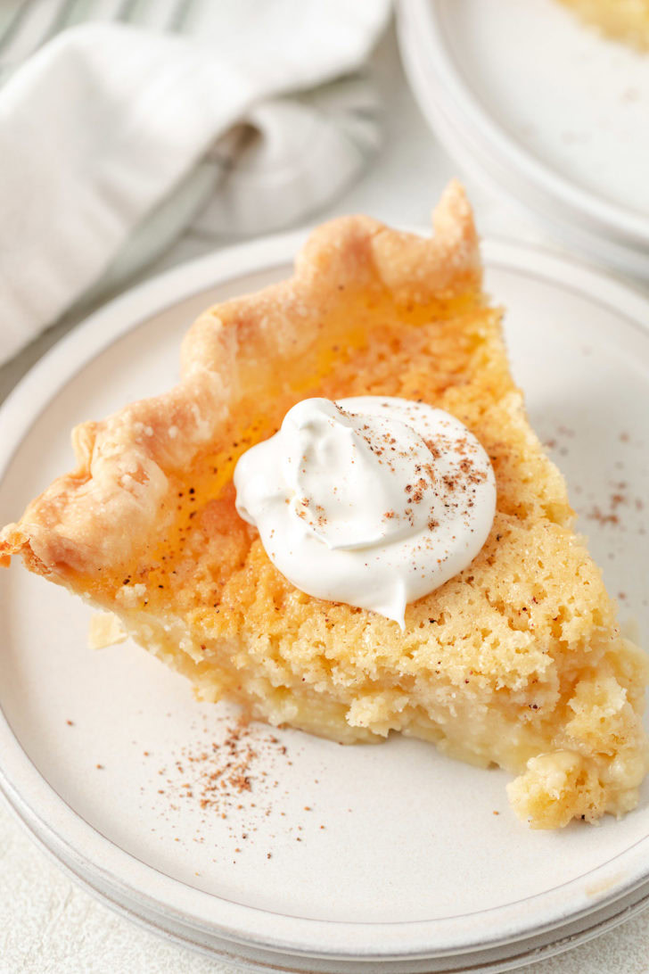 Slice of buttermilk pie on a stack of plates, finished with whipped cream and a dusting of nutmeg.