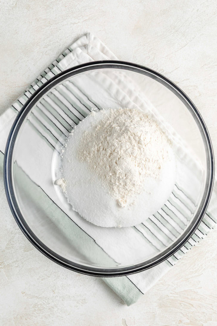 Glass mixing bowl filled with sugar and flour sitting on a striped kitchen towel.