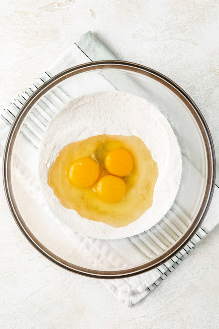 Three raw eggs added to the center of a bowl of sugar and flour.