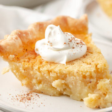 Slice of buttermilk pie topped with whipped cream and a sprinkle of nutmeg on a white plate.