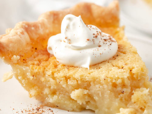 Slice of buttermilk pie topped with whipped cream and a sprinkle of nutmeg on a white plate.