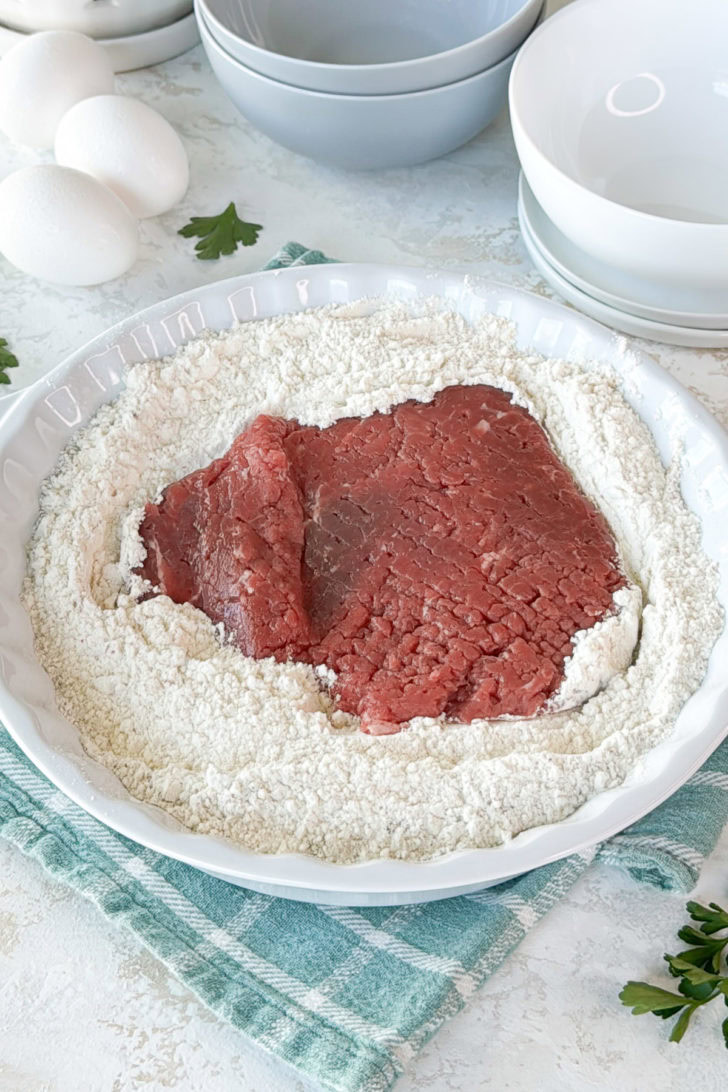 Cube steak sitting in a dish of seasoned flour mixture.
