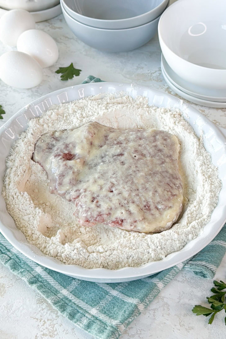 Cube steak being dipped into seasoned flour.