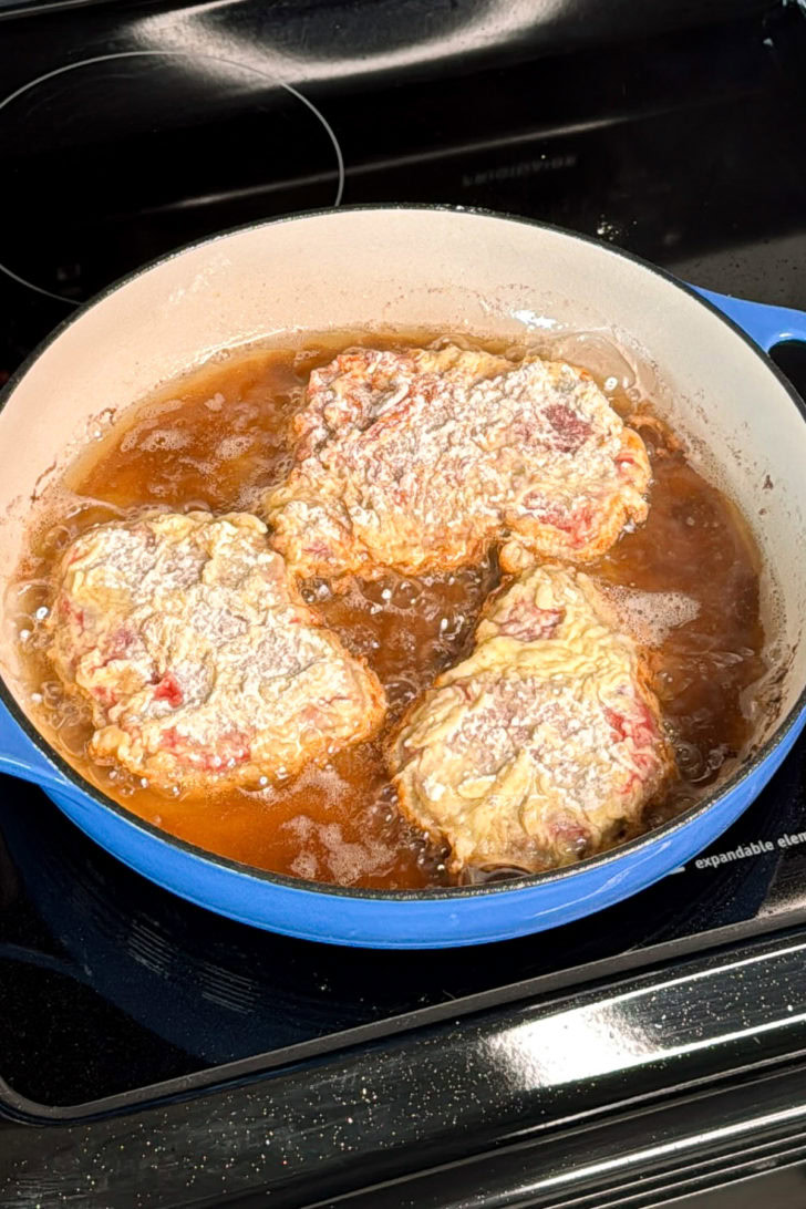 Chicken fried steak pieces frying in hot oil in a skillet on the stovetop.