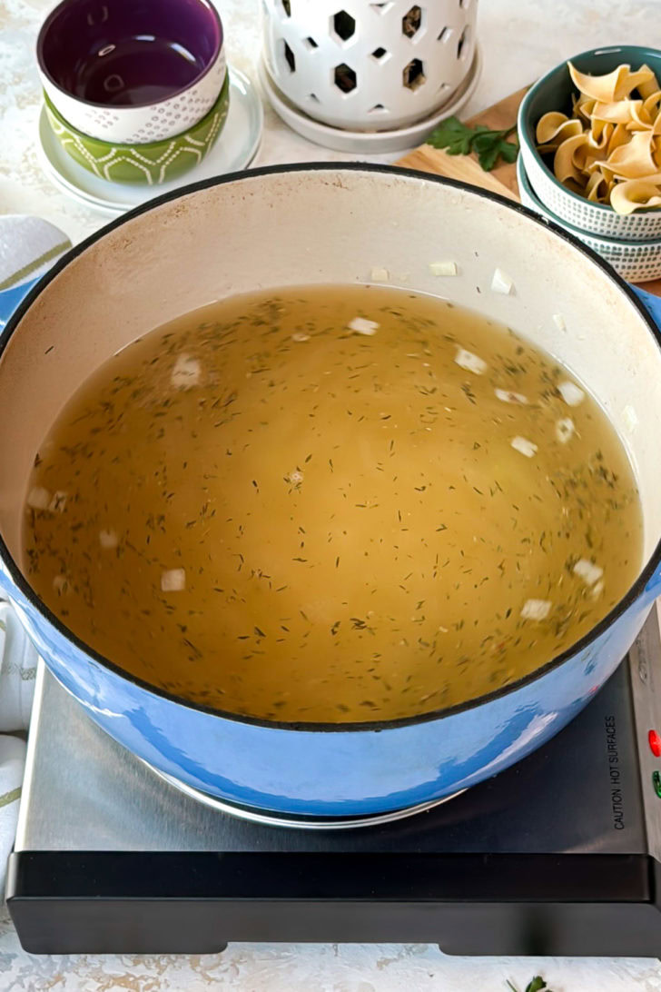 Creamy chicken noodle soup broth base in a blue Dutch oven with chicken broth and dried herbs before adding noodles and chicken.