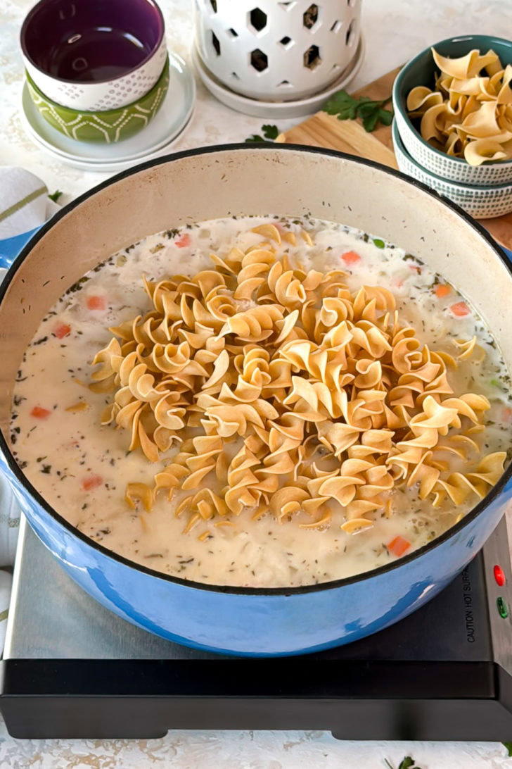Dry egg noodles added to a pot of creamy soup base in a blue Dutch oven.
