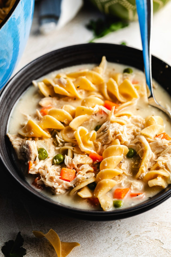 Close-up of chicken noodle soup in a black bowl with wide egg noodles, shredded chicken, peas, and diced carrots.