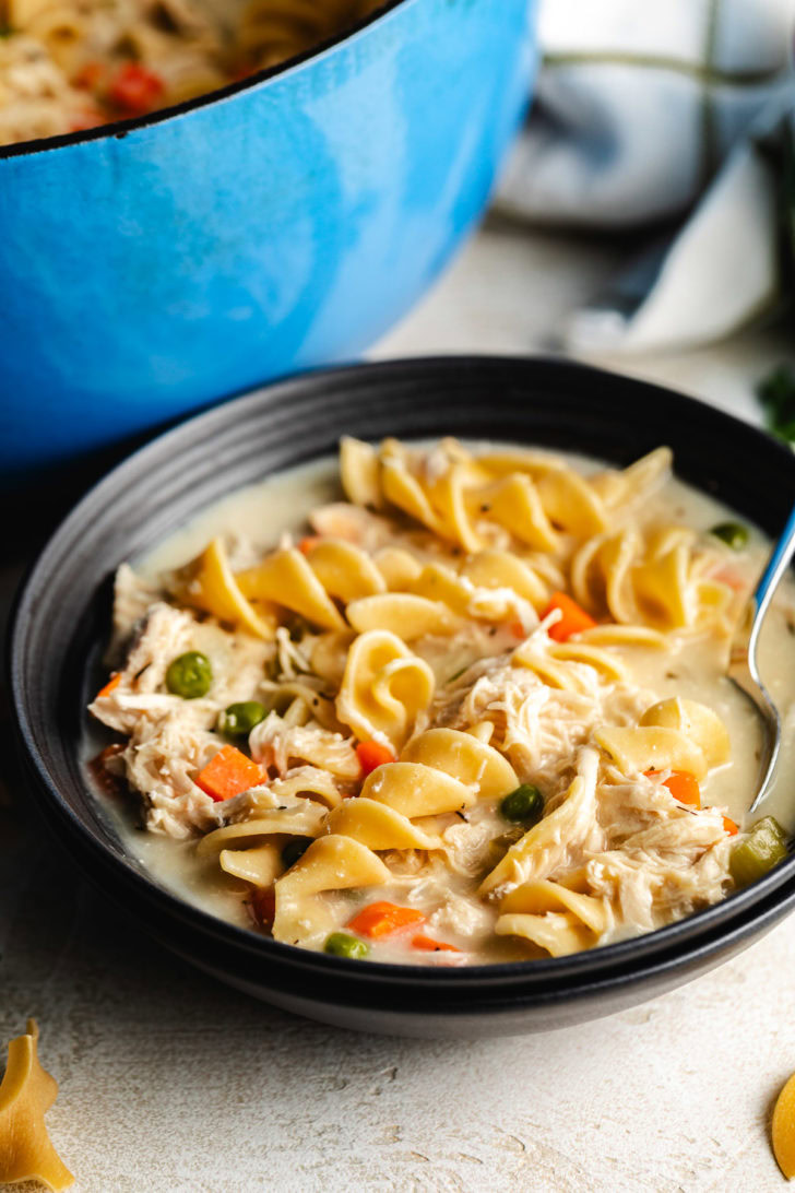 Creamy chicken noodle soup in a black bowl with egg noodles, shredded chicken, peas, and carrots, with a blue pot in the background.