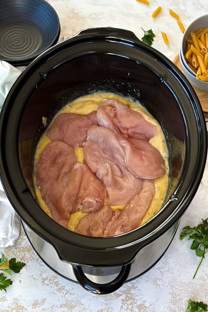 Top-down photo of raw chicken breasts sitting in a creamy seasoned mixture inside a slow cooker.