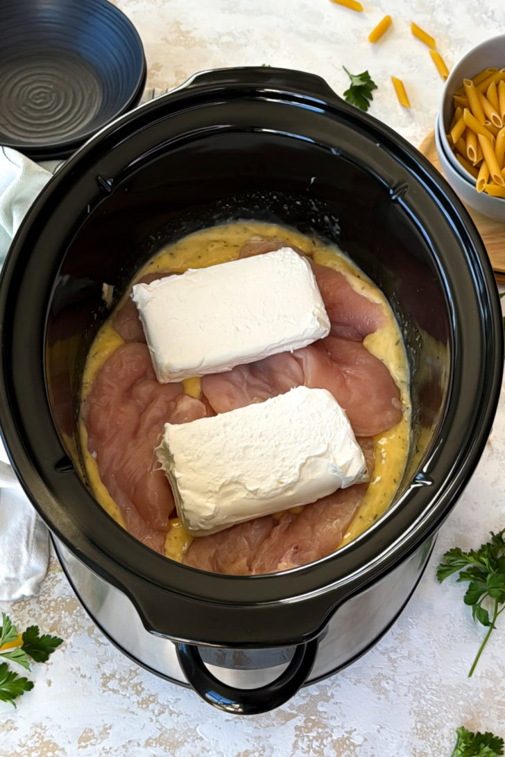 Crockpot Italian chicken pasta prep in a slow cooker with raw chicken breasts and two blocks of cream cheese.