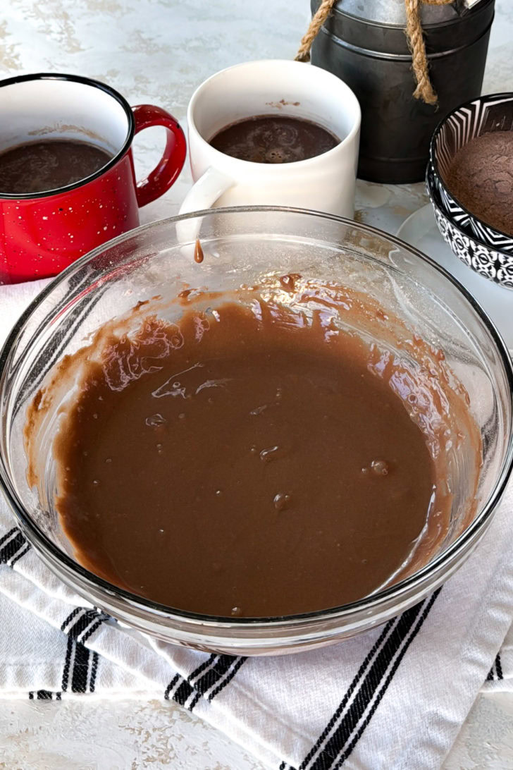 Smooth chocolate filling in a glass mixing bowl, with mugs of cocoa and a chocolate crust in the background.