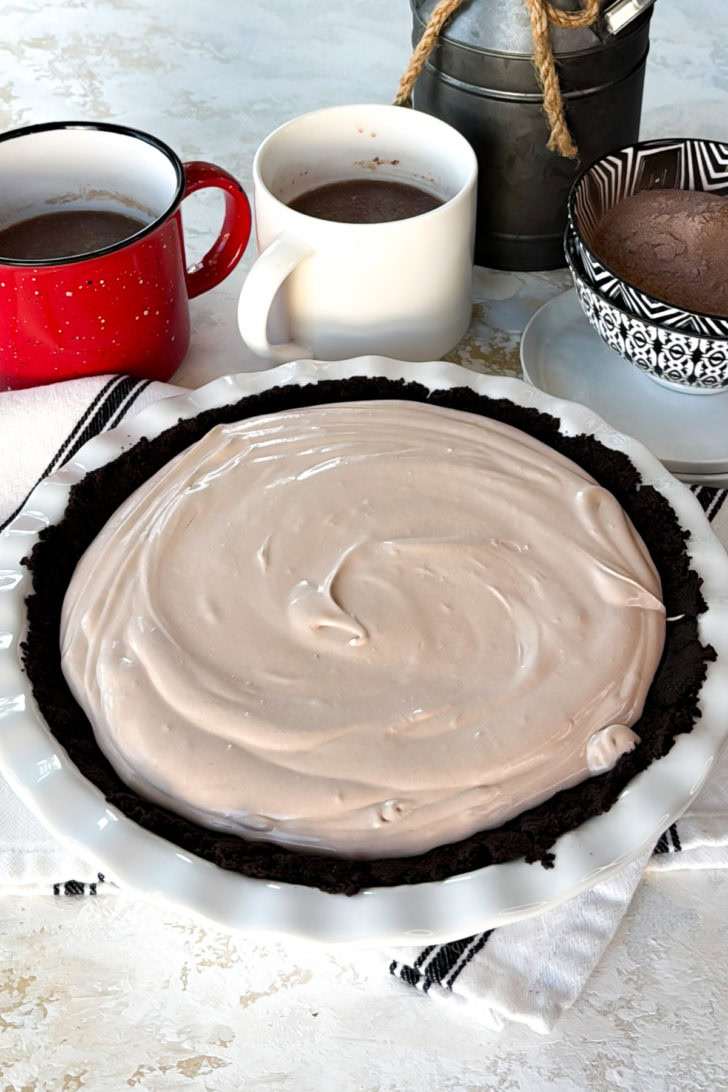 Chocolate mousse filling smoothed into a premade chocolate cookie crust, with mugs of cocoa and hot chocolate mix nearby.