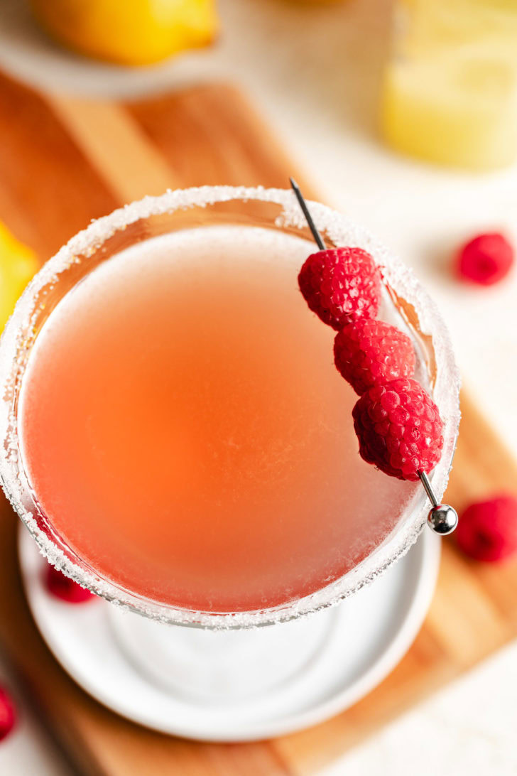 Top-down view of a pink cocktail in a sugar-rimmed martini glass with a fresh raspberry skewer garnish.