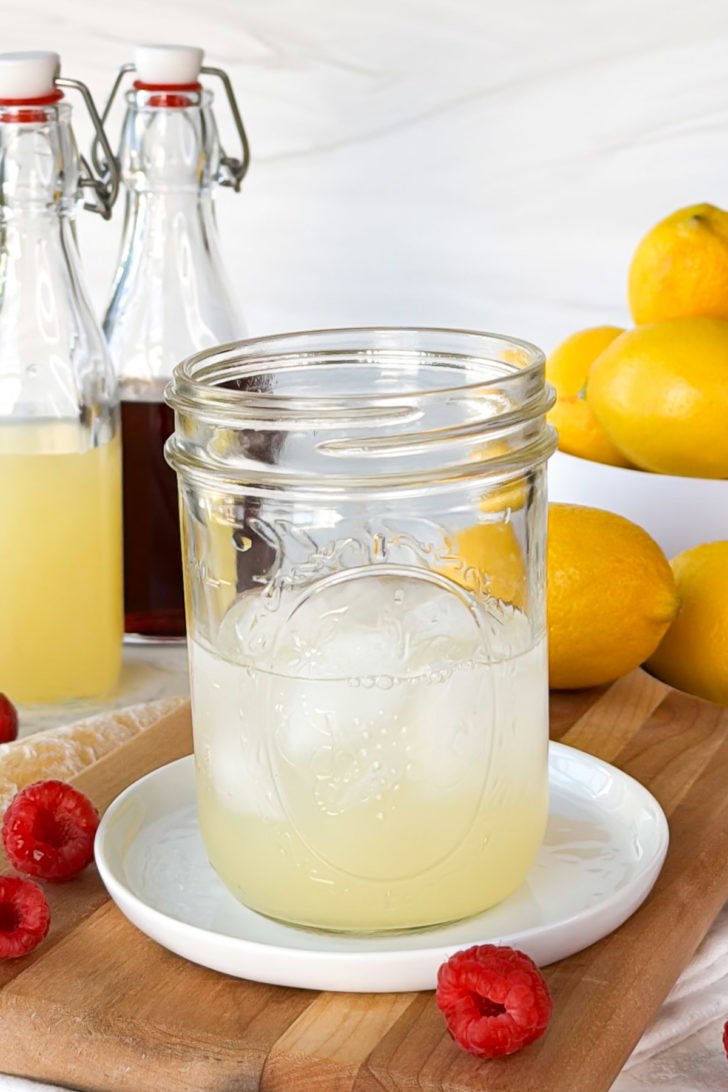 Lemon juice mixture in a mason jar over ice, staged with fresh raspberries and whole lemons on a wooden board.