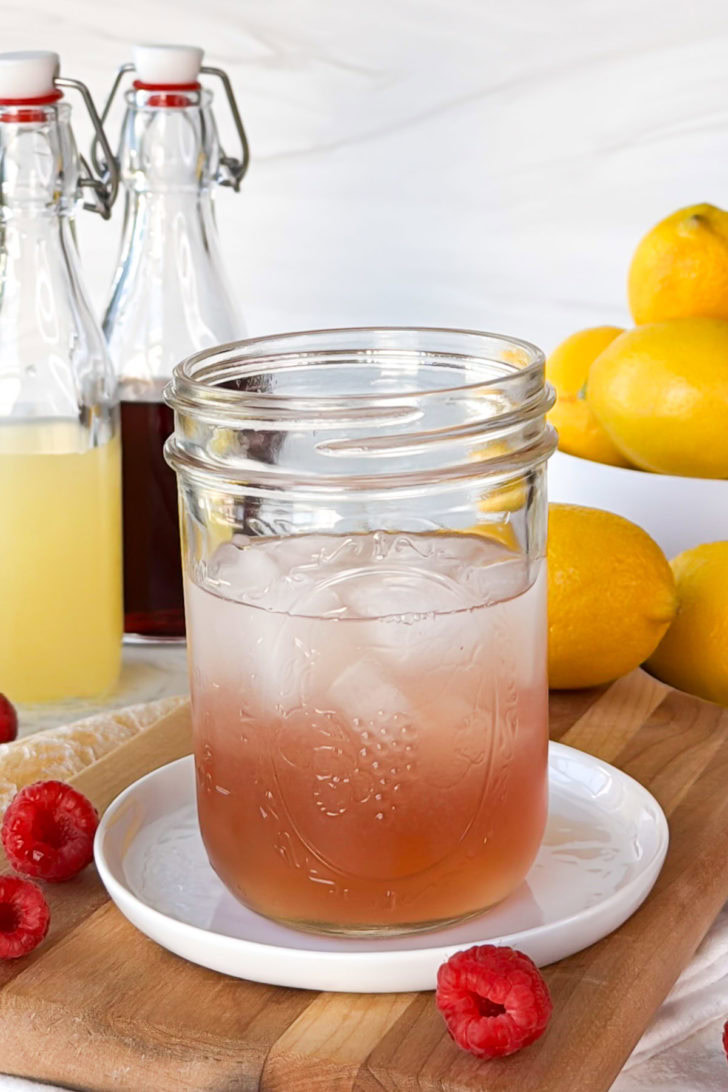 Pink vodka cocktail mixture in a mason jar with ice, photographed with raspberries, lemons, and simple syrup bottles in the background.