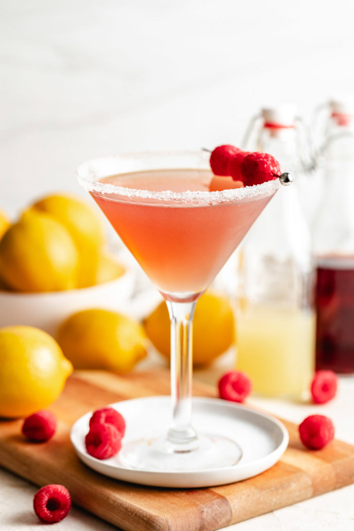 Martini glass with a sugared rim and pale pink drink, styled with raspberries, lemon juice, and simple syrup bottles behind it.