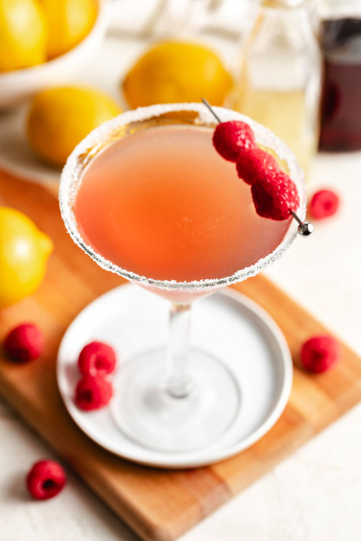 Top view of a raspberry lemon drop martini with a sugared rim and raspberry skewer garnish, with lemons and bottles softly blurred behind.
