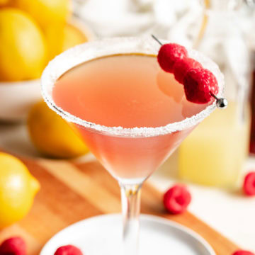 Raspberry lemon drop martini in a sugar-rimmed martini glass with a fresh raspberry garnish and lemons in the background.
