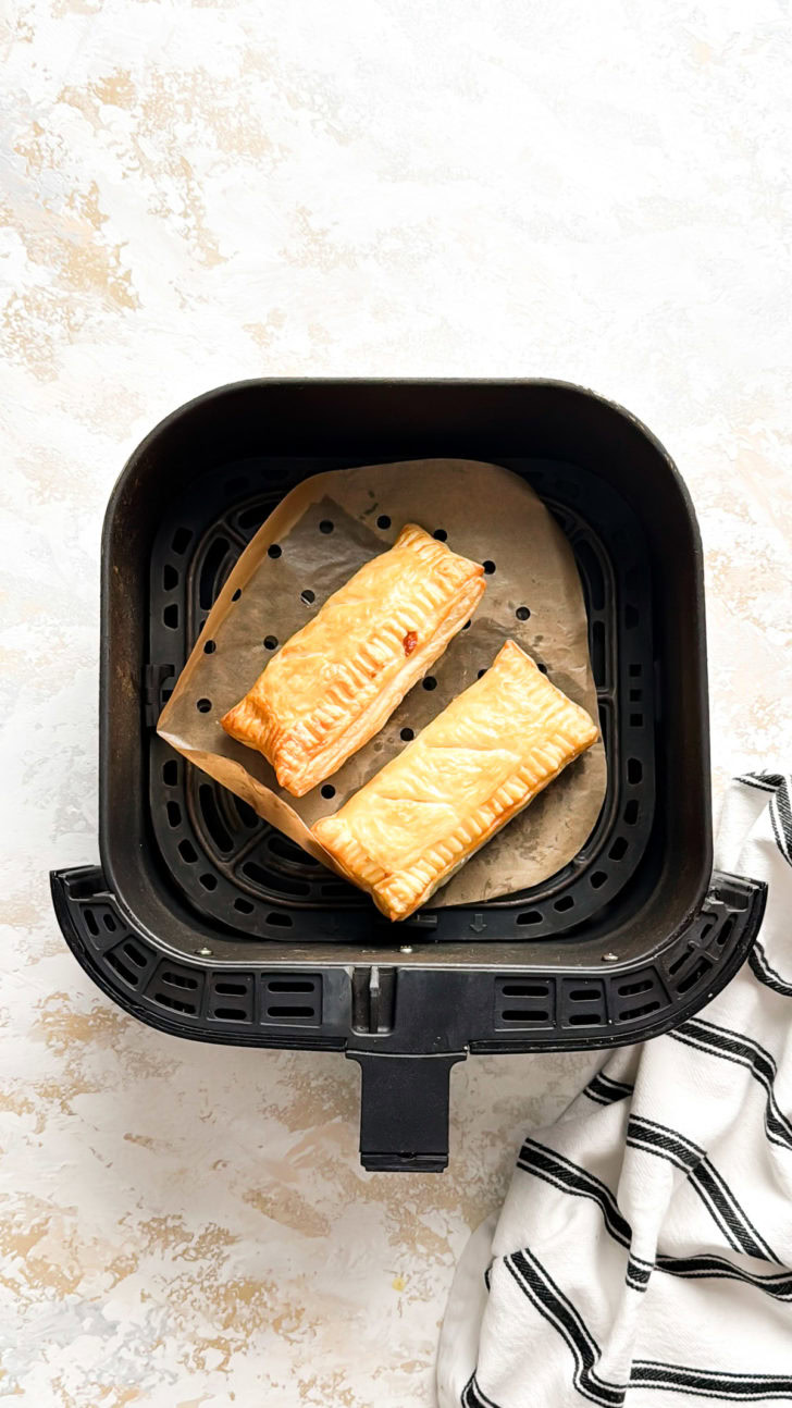Two golden brown puff pastry pizza pockets resting on parchment in the air fryer basket after cooking.