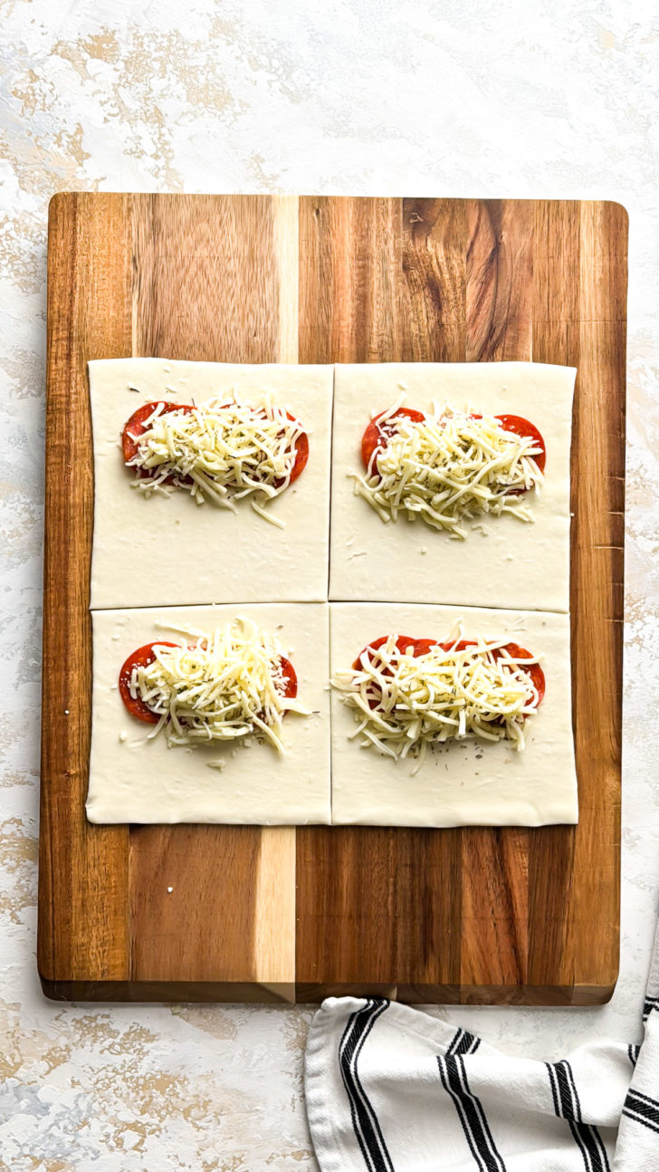 Unbaked puff pastry squares on a wooden board topped with sliced pepperoni, shredded mozzarella, and Italian seasoning ready to be folded into pizza pockets.