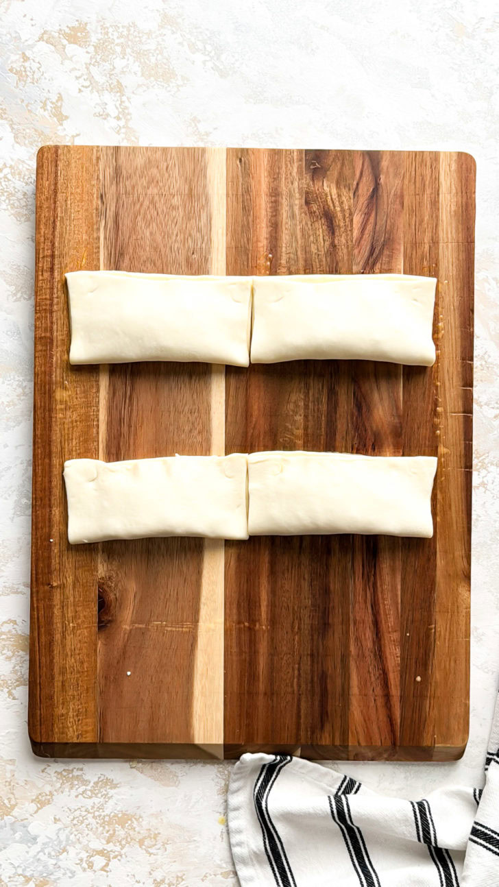 Four folded puff pastry logs filled with pepperoni and cheese lined up on a wooden board before crimping and cooking.