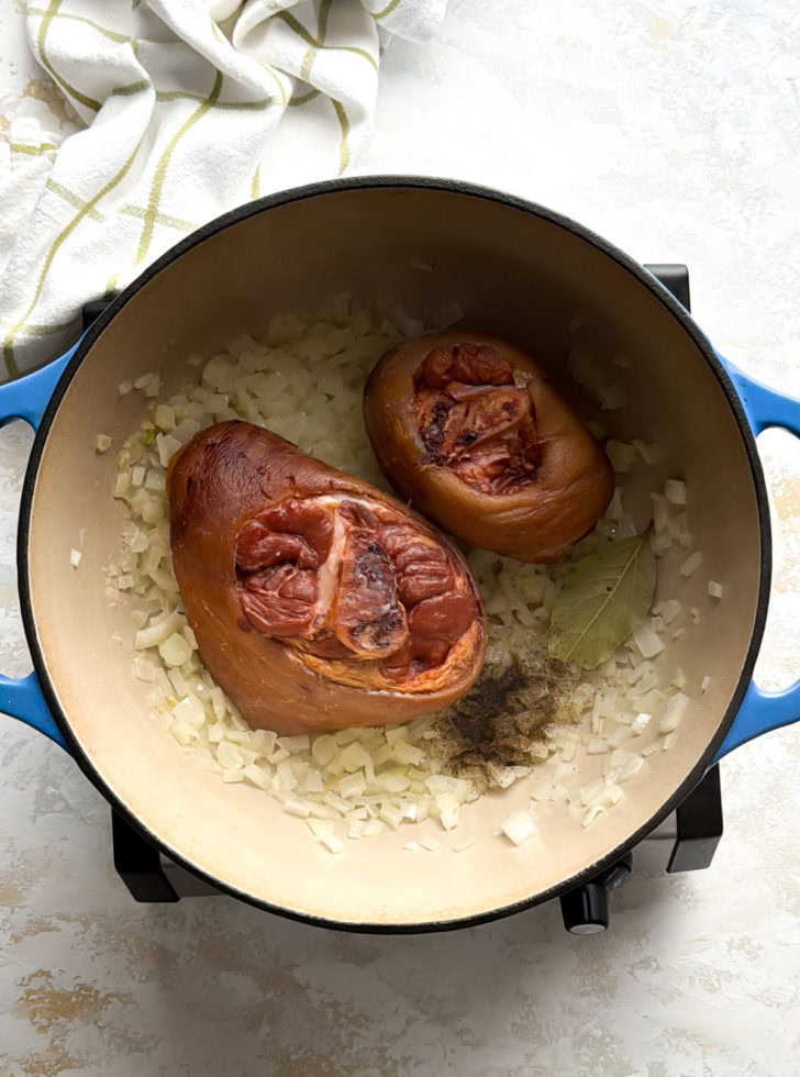 Two smoked ham hocks with onions, bay leaf, and pepper in a pot.