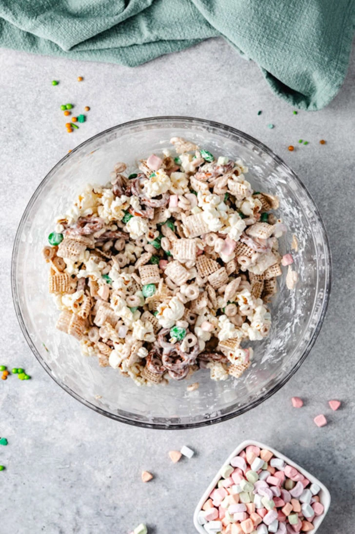 Snack mix tossed together in a glass bowl with popcorn, cereal, pretzels, marshmallows, and green chocolate candies.