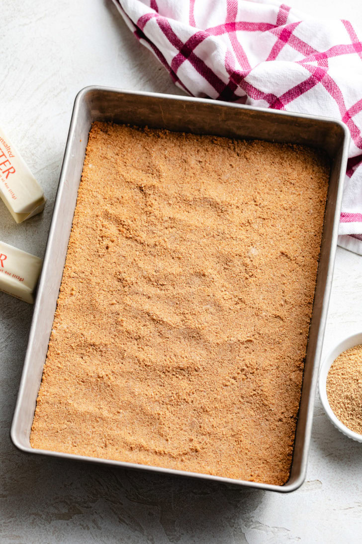 A golden brown crumb base is firmly packed into a metal baking dish next to a pink plaid towel.