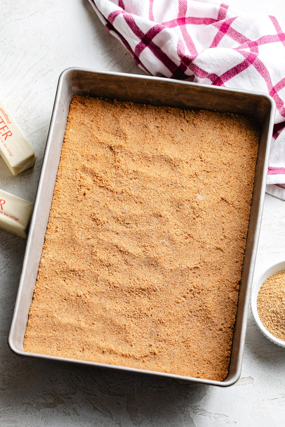 A golden brown crumb base is firmly packed into a metal baking dish next to a pink plaid towel.