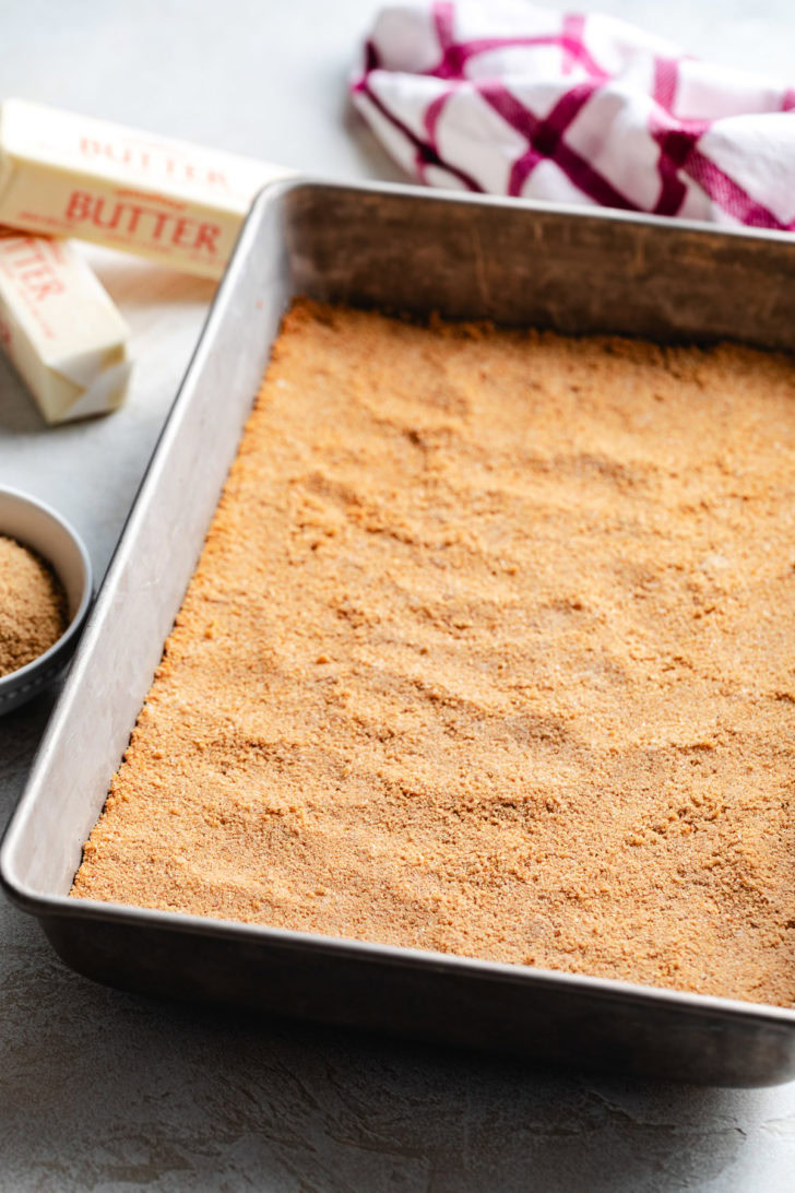 A sweet graham cracker crust is pressed evenly into a large rectangular baking pan.
