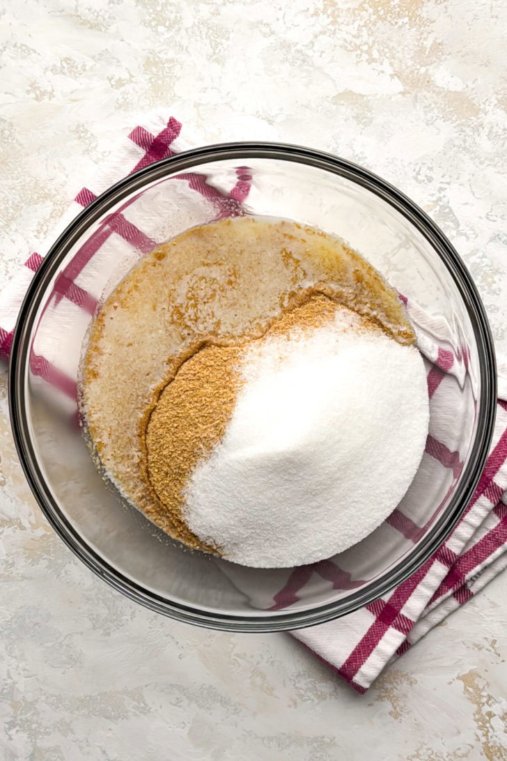 Granulated sugar, dry crumbs, and melted butter sit unmixed in a clear glass bowl.