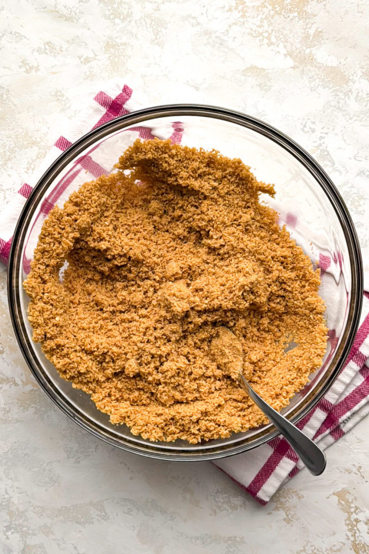 A spoon rests in a glass bowl after stirring the buttery crumb mixture until it resembles wet sand.