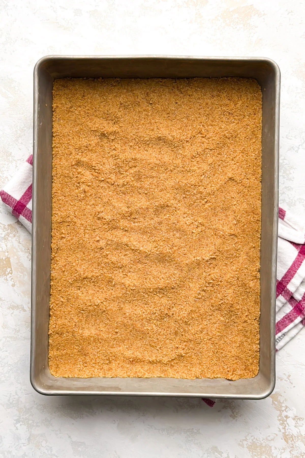 An overhead view shows a perfectly even graham cracker crust covering the bottom of a pan.