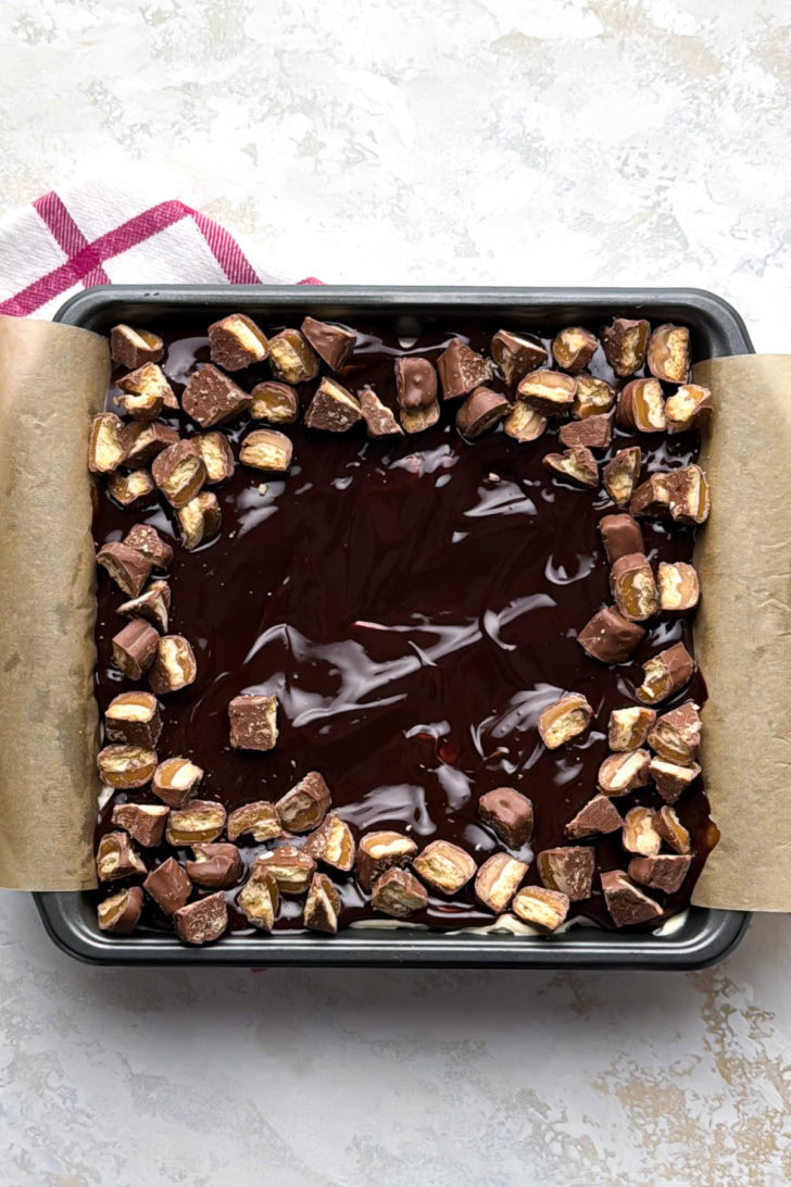 Finished dessert bars in a pan with ganache and chopped candy bars.