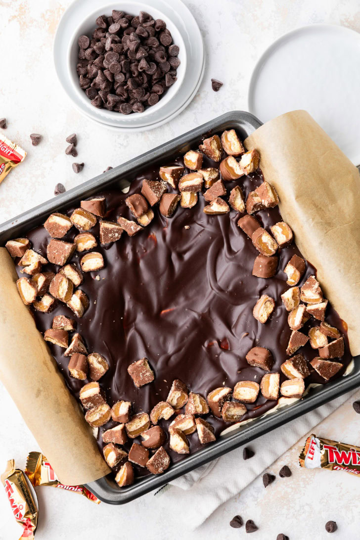 Twix cheesecake bars in a square pan with a glossy chocolate topping.
