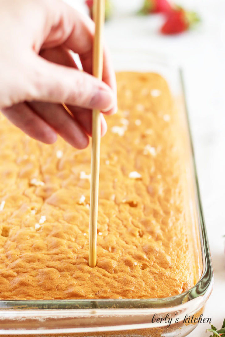Poking holes in baked tres leches cake with a wooden skewer.